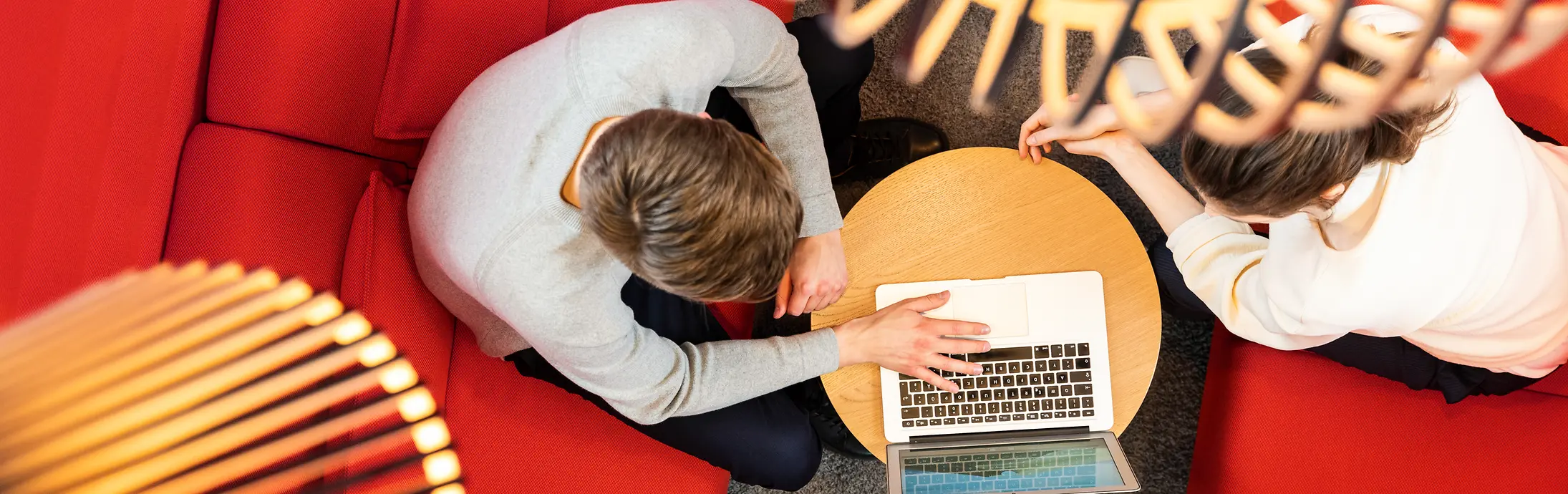 Duas pessoas sentadas em um sofá vermelho e trabalhando juntas em um laptop 