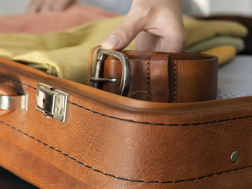 Imagem de uma pessoa arrumando uma mala de couro marrom com fivela e um cinto e roupas