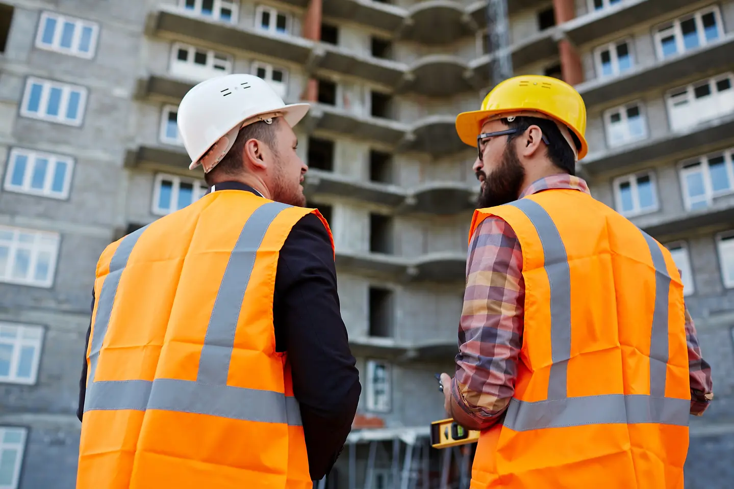 Imagem de dois profissionais da construção uniformizados conversando celular em frente a um prédio