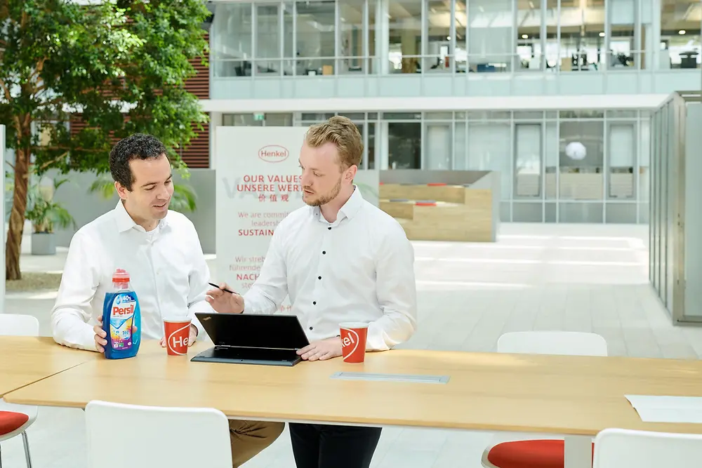 Dois homens sentados em uma mesa com um frasco de Persil e um laptop