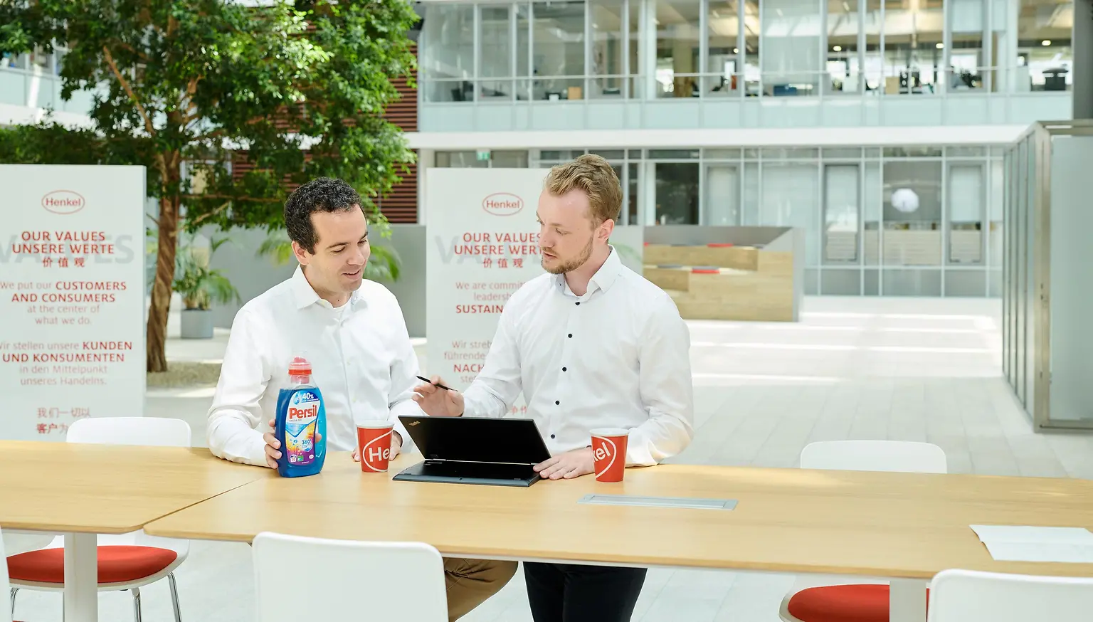 Dois homens sentados em uma mesa com um frasco de Persil e um laptop